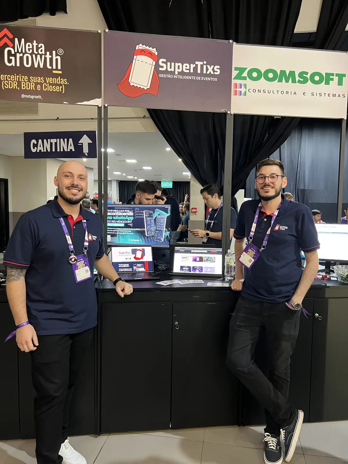 Dois integrantes da equipe SuperTixs no estande da marca no Empreende SC, com notebooks exibindo a plataforma